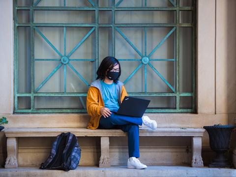 A young man wearing a mask sitting on a stone bench in front of windows with an open laptop.